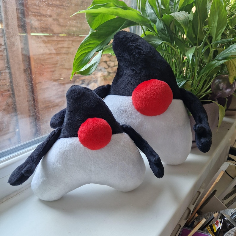 May include: Two plush toys shaped like a penguin with black and white bodies and red beaks. The larger penguin is standing upright, while the smaller penguin is lying down.
