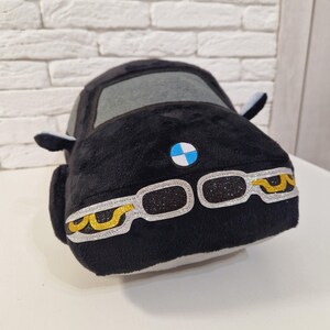 以下が含まれることがあります： 青と白のBMWロゴと金のアクセントが付いた黒いぬいぐるみ車。車は灰色のフロントガラスと白いインテリアです。