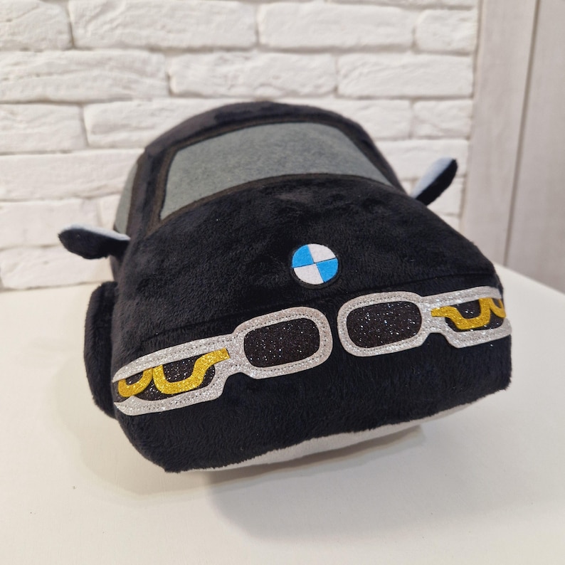 以下が含まれることがあります： 青と白のBMWロゴと金のアクセントが付いた黒いぬいぐるみ車。車は灰色のフロントガラスと白いインテリアです。