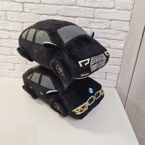 以下が含まれることがあります： 黒のぬいぐるみ製の車が2台、積み重ねられています。上の車はグレーのインテリアと、シルバーのグリルに「Audi」と書かれています。下の車は、フロントに青と白のロゴと、黄色のヘッドライトが付いています。