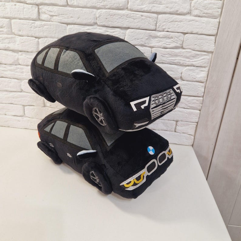 以下が含まれることがあります： 黒のぬいぐるみ製の車が2台、積み重ねられています。上の車はグレーのインテリアと、シルバーのグリルに「Audi」と書かれています。下の車は、フロントに青と白のロゴと、黄色のヘッドライトが付いています。