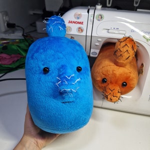 May include: Two plush toys shaped like cacti, one blue and one orange, with embroidered details. The blue cactus has a white nose and black eyes. The orange cactus has a black nose and black eyes. The toys are sitting on a white surface.
