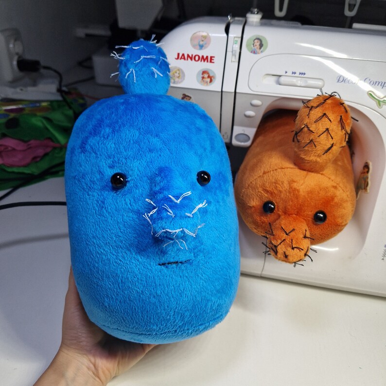 May include: Two plush toys shaped like cacti, one blue and one orange, with embroidered details. The blue cactus has a white nose and black eyes. The orange cactus has a black nose and black eyes. The toys are sitting on a white surface.