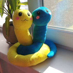 Pu&ograve; includere: Un giocattolo di peluche a forma di serpente giallo e blu con un viso sorridente. Il serpente &egrave; arrotolato in un cerchio.