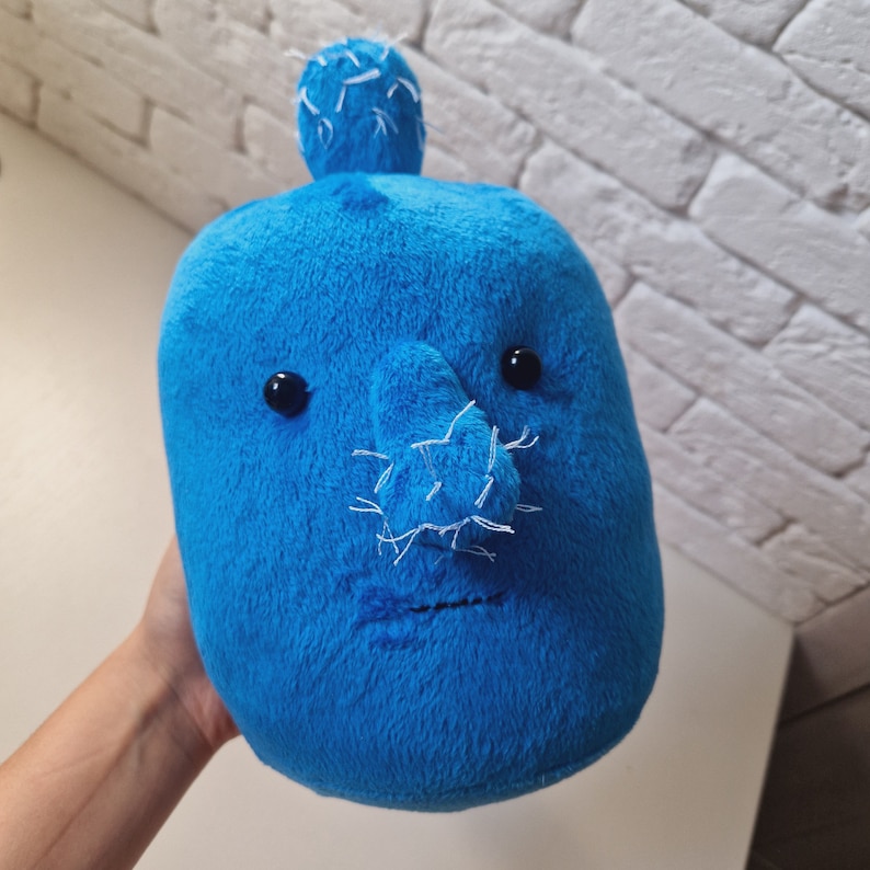 May include: A blue plush toy with a stitched-on face, featuring a large nose, two eyes, and a stitched-on mouth. The toy has a spiky, white, stitched-on hair tuft on top of its head.