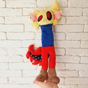 May include: A plush toy with a yellow head, red pants, blue shirt, and a red tail with black spots. The toy has large red eyes and pink ears.