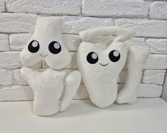 Puede incluir: Cuatro juguetes de peluche blancos con forma de hueso con caras sonrientes y ojos negros. Los juguetes están colocados sobre una superficie blanca contra una pared de ladrillos blancos. Los juguetes incluyen un fémur, una escápula y otras dos formas de hueso.