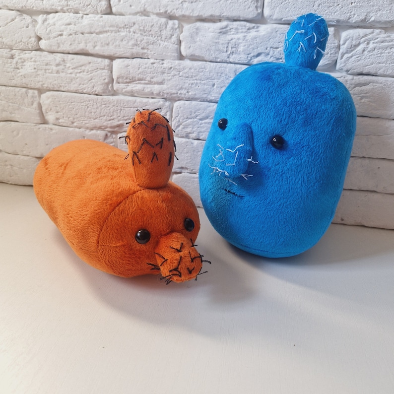 May include: Two plush cactus toys, one blue and one orange. The blue cactus has a face with a stitched-on nose and eyes. The orange cactus has a face with black eyes and a stitched-on mouth.