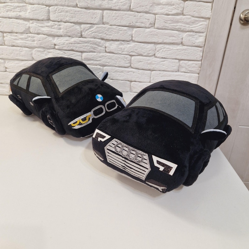 以下が含まれることがあります： 黒いぬいぐるみ製の車2台。1台はフロントに青と白のロゴ、もう1台は銀色のロゴが付いています。両方の車に灰色の窓が付いています。
