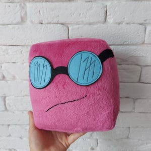 May include: A plush, pink cube-shaped pillow with blue-tinted glasses and a black frame. The glasses have a white design inside. A black, curved line forms a mouth. The pillow is held in a hand.