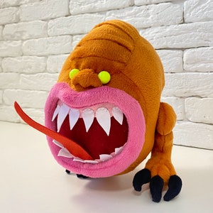 May include: A plush toy monster with orange fur, a large pink mouth with white teeth, and a red tongue sticking out. The monster has green eyes and a menacing expression.