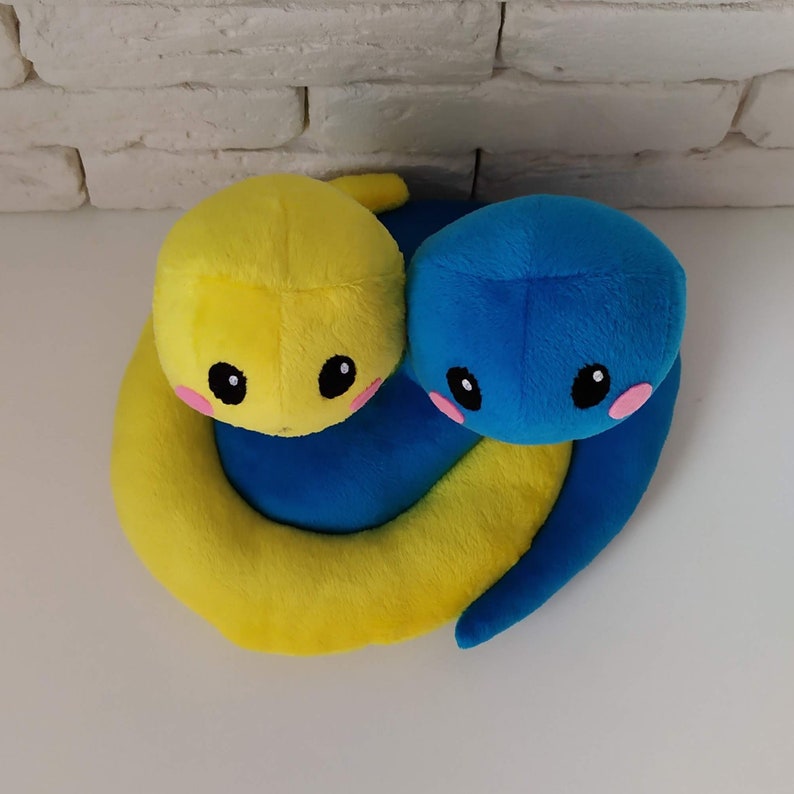 Pu&ograve; includere: Due giocattoli di peluche a forma di serpente, uno giallo e uno blu, sono avvolti l'uno intorno all'altro. Hanno volti carini e da cartone animato con occhi neri e guance rosa.