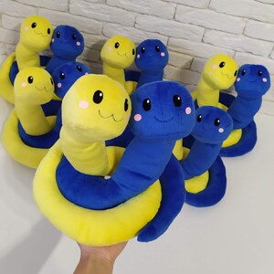 Pu&ograve; includere: Un gruppo di giocattoli di peluche a forma di serpente di colore giallo e blu. I serpenti sono arrotolati in una posa giocosa. Alcuni dei serpenti hanno facce sorridenti con guance rosa.