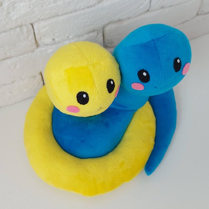 Pu&ograve; includere: Un giocattolo di peluche a forma di serpente giallo e blu con un viso carino. Il serpente &egrave; arrotolato in un cerchio con la testa appoggiata sulla coda.