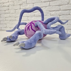 Op de afbeelding: Een pluche speelgoedbeest met een ronde, lichtpaarse body en meerdere blauwe aanhangsels. De body heeft een donkerder paars, aderachtig design. Het beest heeft meerdere blauwe poten met grijze klauwen en verschillende gebogen, blauwe ranken.