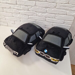 以下が含まれることがあります： 黒いぬいぐるみ製の車のクッション2つ。1つは灰色のフロントガラスと白いヘッドライトが付いた黒いアウディ。もう1つは灰色のフロントガラス、白いヘッドライト、青いBMWロゴが付いた黒いBMWです。