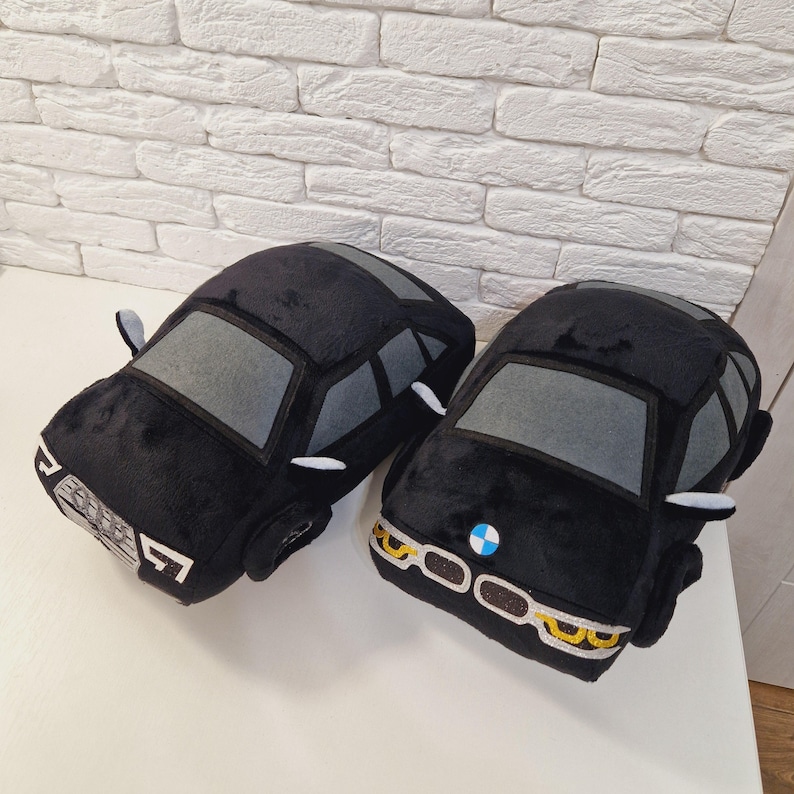 以下が含まれることがあります： 黒いぬいぐるみ製の車のクッション2つ。1つは灰色のフロントガラスと白いヘッドライトが付いた黒いアウディ。もう1つは灰色のフロントガラス、白いヘッドライト、青いBMWロゴが付いた黒いBMWです。