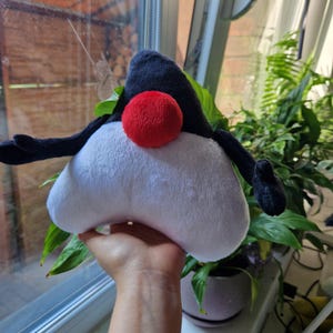 May include: A white and black plush toy shaped like a triangle with a red nose. The toy has long arms and a black hat.