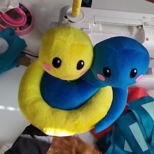 Pu&ograve; includere: Un giocattolo di peluche giallo e blu a forma di serpente. Il serpente &egrave; arrotolato su se stesso con la testa appoggiata sulla coda. Il serpente ha grandi occhi neri e guance rosa.