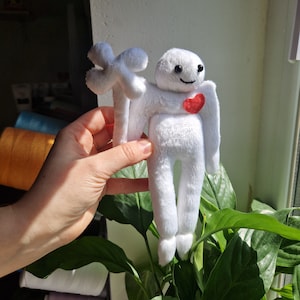 Puede incluir: Juguete de peluche blanco con un corazón rojo en el pecho y una flor blanca en la mano. El juguete sonríe y tiene una expresión amigable.