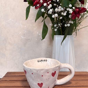 May include: A speckled ceramic mug with red heart designs and the word "CARIAD" on the rim. The mug is on a wooden surface, with a white vase holding red flowers and white baby's breath in the background.