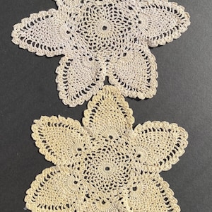 Puede incluir: Dos tapetes de ganchillo hechos a mano, de color crema, con un diseño floral. Los tapetes tienen forma de seis pétalos con un centro calado detallado y bordes festoneados. La textura del ganchillo es visible, mostrando la artesanía.