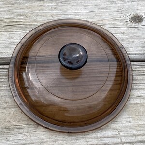 May include: A Pyrex V-1-B brown glass lid with a dark brown knob. The lid is round and transparent, with a slightly raised centre and a visible rim. The lid is designed for cookware.