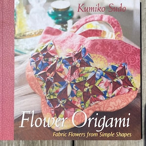 May include: A book titled "Flower Origami" by Kumiko Sudo. The cover features a close-up of a colorful fabric flower arrangement. The book's spine is pink with a dotted pattern, and the background is a wooden surface.