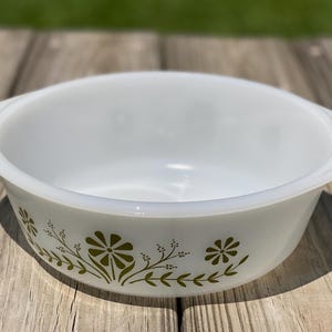May include: A white milk glass casserole dish with integrated side handles. The exterior features a repeating pattern of olive green stylized flowers, leaves, and delicate sprigs encircling the bowl. The interior is smooth, white, and glossy.