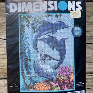 May include: A Dimensions counted cross stitch kit featuring three blue dolphins swimming in an underwater scene. The kit includes cotton thread, a 14-count blue Aida cloth, a needle, and instructions. The finished design measures 5" x 7".
