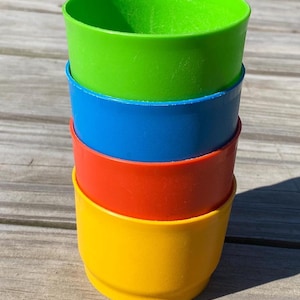May include: A stack of four cylindrical plastic cups in bright solid colors. The top cup is lime green, followed by a blue cup, an orange cup, and a yellow cup at the base. Each cup features a smooth, matte finish and a simple, open-top design.