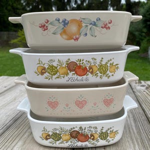 May include: Four rectangular ceramic baking dishes stacked. Each dish has white handles and is decorated with fruit and vegetable designs. The top dish has a fruit design, the second and fourth dishes have a vegetable design, and the third dish has a heart design.