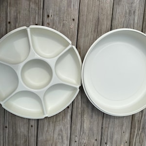Puede incluir: Una bandeja de servicio redonda de plástico color blanco hueso que presenta un compartimento circular central rodeado por cinco secciones distintas en forma de cuña, acompañada de una tapa redonda a juego con superficie interior lisa.