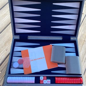 May include: Open backgammon set in a gray case with a blue interior. The set includes white and red game pieces, dice, and an orange booklet labeled "BACKGAMMON". The case is open, revealing the game board and accessories.