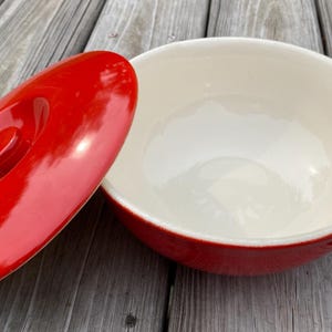 May include: A red ceramic casserole dish with a matching lid. The dish has a white interior and a red exterior. The lid is oval-shaped with a central handle. The dish is designed for cooking and serving.