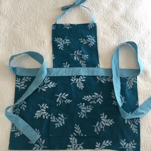 May include: A teal blue apron with a white leaf pattern and a light blue trim. The apron has a large pocket in the front and adjustable straps.