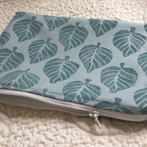 May include: A light blue zippered pouch featuring a repeating pattern of teal-colored leaves. The leaves are stylized and printed on the fabric. The zipper is white, and the pouch is resting on a white, quilted surface.