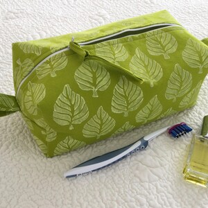 Puede incluir: Un neceser verde lima con un estampado de hojas blancas, una cremallera blanca y un asa pequeña. Un cepillo de dientes y un frasco de perfume están junto a la bolsa.