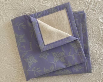May include: A lavender-colored fabric napkin with a white floral pattern. The napkin is folded in half and has a white cotton lining.