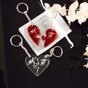 May include: Two red acrylic heart-shaped keychains with a puzzle piece design. The keychains are engraved with the letters "K" and "J" and small hearts. The keychains are attached to silver chains and rings.