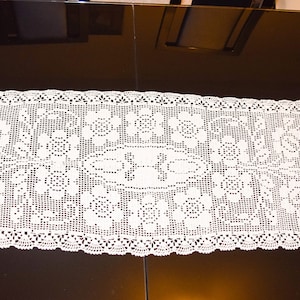 Puede incluir: Un camino de mesa blanco de ganchillo con un diseño floral. El camino de mesa tiene un borde festoneado y está hecho de encaje delicado.