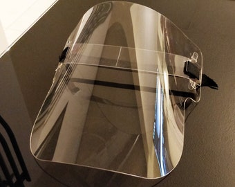 May include: A clear plastic face shield with a black elastic band. The shield is curved and has a wide field of view.