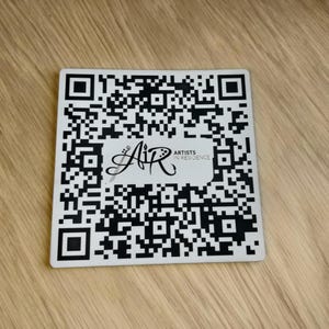 May include: A white square with a black and white QR code. The QR code is surrounded by a black border. In the center of the QR code is a black and white logo that says "AIR" with the words "ARTISTS IN RESIDENCE" below it.