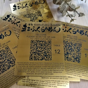 Pode incluir: Sinais de número de mesa dourados com um código QR e o texto "Bem-vindo, Digitalize o código QR para ver nosso menu" em preto. O texto "Marufuku Ramen" também está em preto e apresenta os caracteres japoneses "ふっくらあめん". Os números de mesa são 5, 12 e 1.