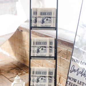 Op de afbeelding: Een zwarte metalen krantenrek met meerdere planken, met kranten met de kop "BREAKING NEWS!!" en de namen "RYAN & JENNIFER". De bovenkant van het rek leest "The Newlywed Times, PLEASE TAKE ONE".