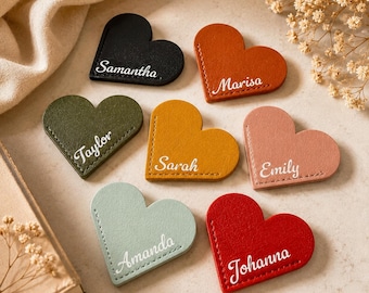 May include: A collection of heart-shaped leather bookmarks in various colors, including black, orange, green, yellow, pink, blue, and red. Each bookmark features a name in white script: Samantha, Marisa, Taylor, Sarah, Emily, Amanda, and Johanna.