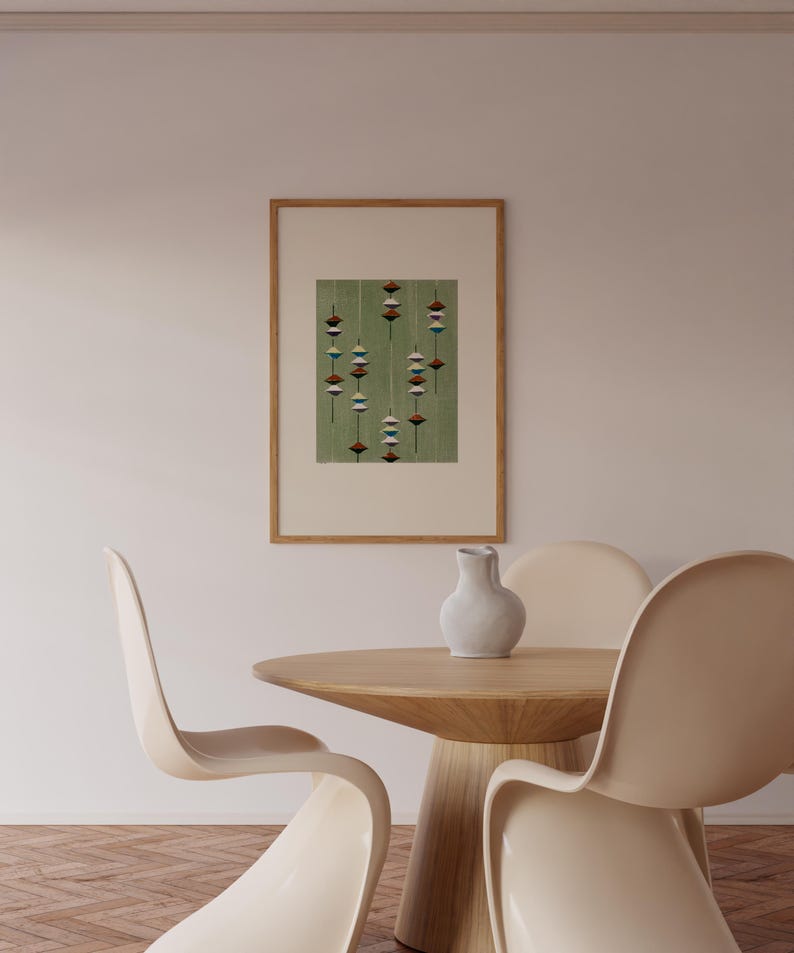 May include: A framed artwork featuring a green background with a pattern of hanging, stylized shapes in shades of red, white, and blue. The artwork is displayed above a wooden table and chairs in a neutral-toned room.