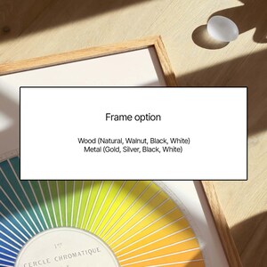 May include: A framed colour wheel print with a natural wood frame. The text "Frame option" is displayed above a list of wood and metal frame colours. A small, oval, translucent object sits on the wooden surface.