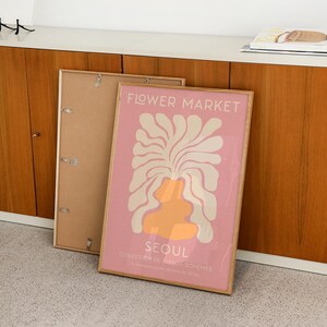 May include: A framed poster with a pink background and a white and orange abstract floral design. The poster reads "FLOWER MARKET SEOUL COLLECTION DE FLEURS BOHEMES 37. GANGNAM-DATRE SEOCHO-GU SEOUL".