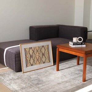 K&ouml;nnte beinhalten: Ein gerahmtes Kunstwerk mit einem geometrischen Muster in Beige-, Burgunder- und Cremet&ouml;nen liegt auf einem grauen Teppich. Das Kunstwerk wird in einem modernen Wohnzimmer pr&auml;sentiert, zusammen mit einem dunkelgrauen Ecksofa und einem Holztisch.